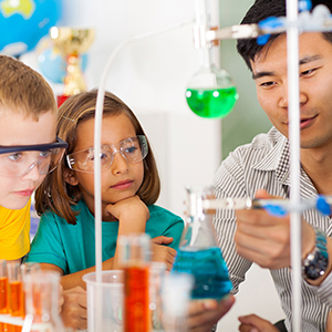 A teacher demonstrating a science experiment with liquid-filled beakers and flasks to two young school students wearing safety goggles.